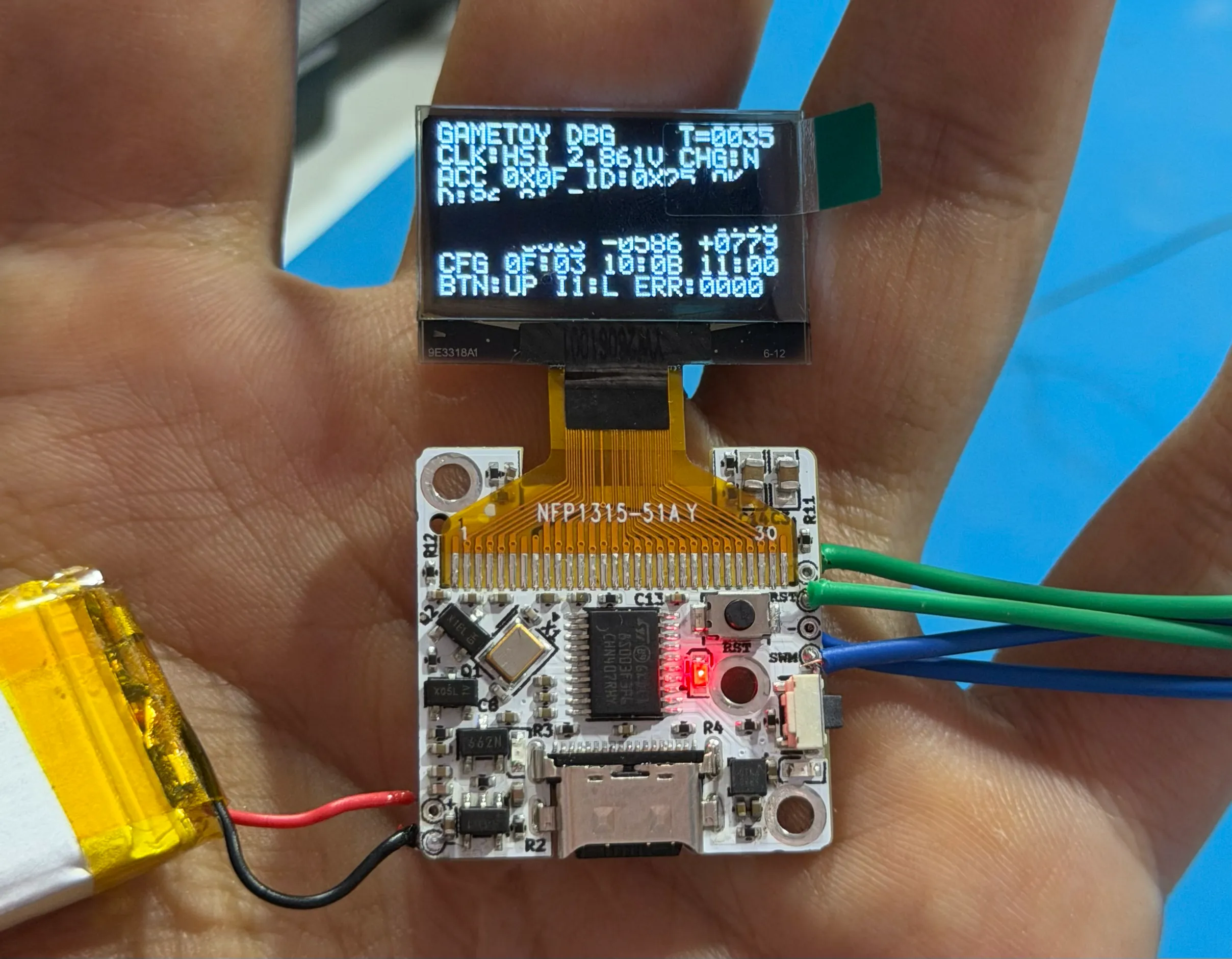 A small white board held in hand, its OLED screen showing boot text, wired to a battery