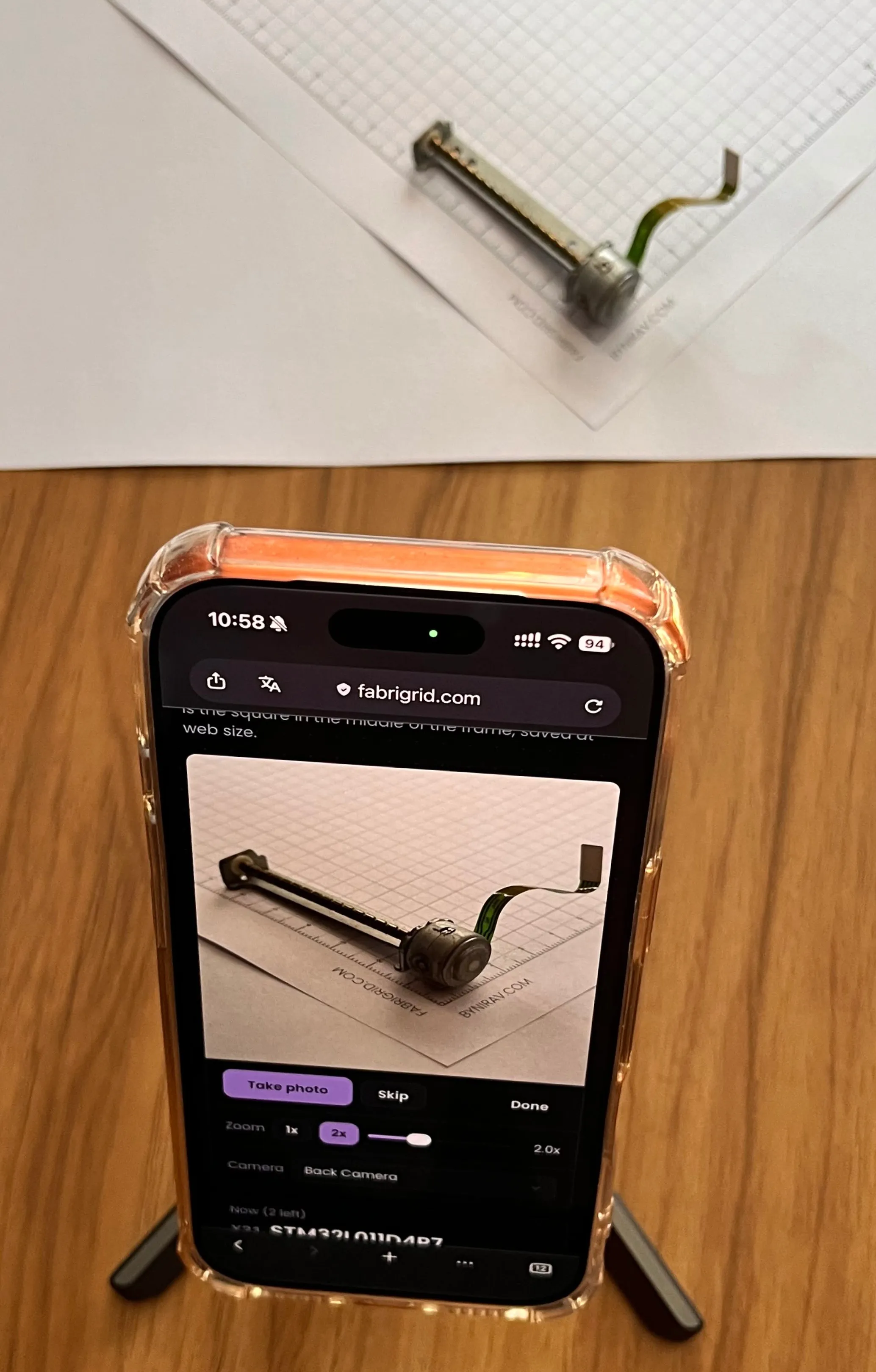 A phone standing on a small tripod, showing fabrigrid.com's photo screen with a stepper motor on graph paper in its viewfinder; the real motor sits on the paper beyond it