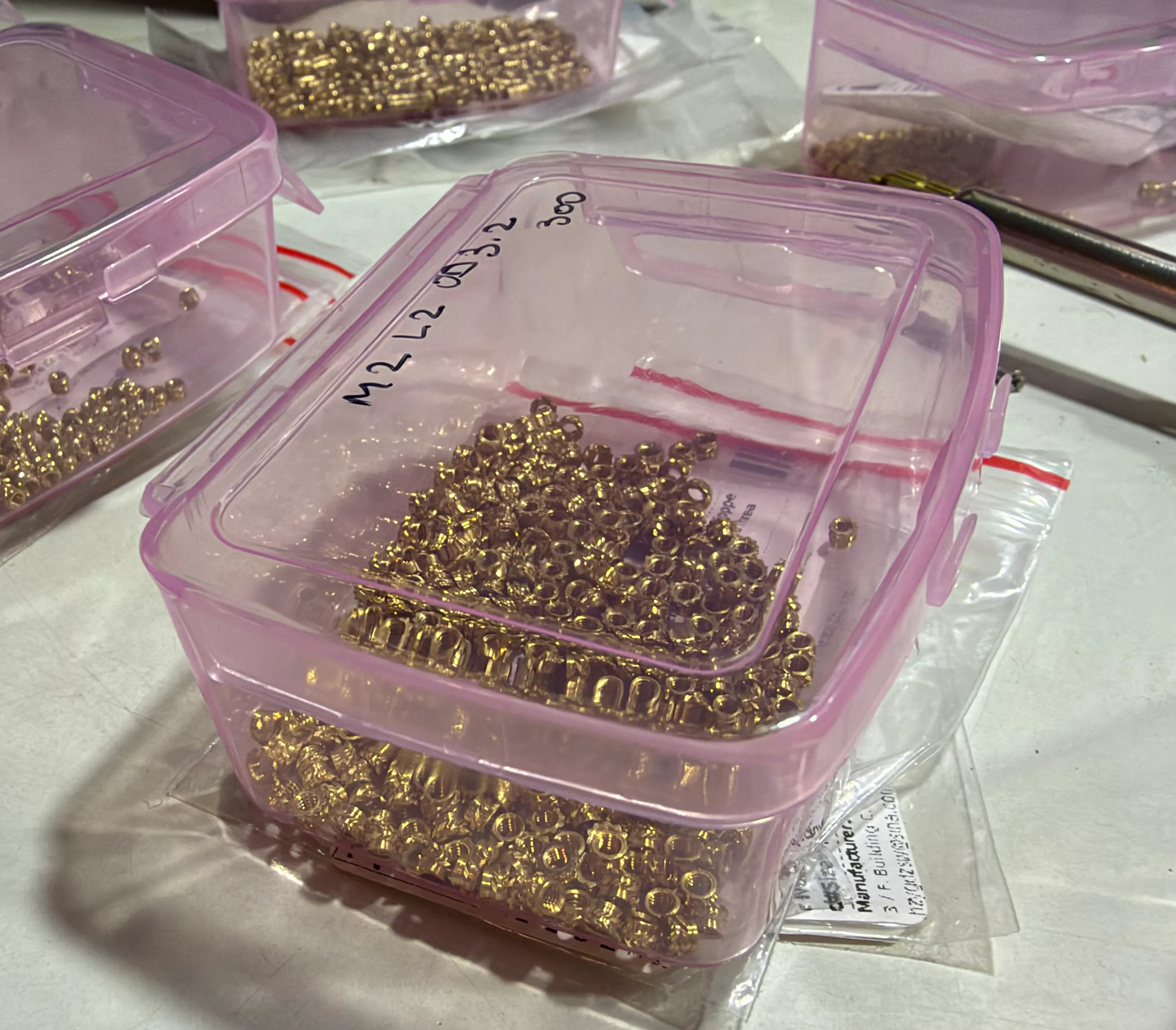 A clear pink plastic box of brass threaded inserts, labelled by hand, with similar boxes behind it